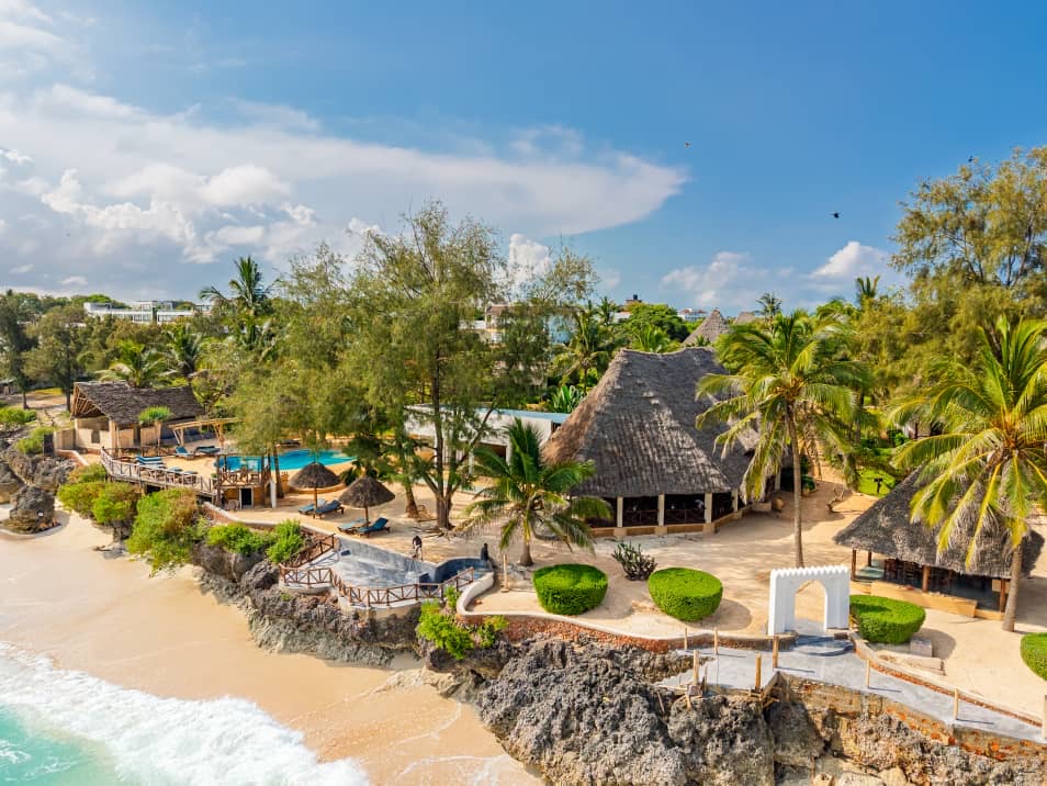 Tanzanite Beach Resort in Nungwi, Zanzibar seen from above — the pool and thatched pavilions on the cliff, the white arch, and the turquoise shore with breaking waves